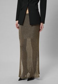 Metallisch goldbrauner Rock mit transparenter Textur, kombiniert mit einem taillierten schwarzen Blazer. Der Rock hat eine lange, fließende Silhouette und Seitenschlitze.