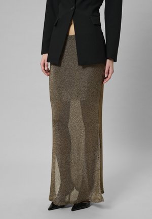Metallisch goldbrauner Rock mit transparenter Textur, kombiniert mit einem taillierten schwarzen Blazer. Der Rock hat eine lange, fließende Silhouette und Seitenschlitze.