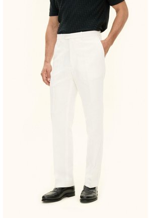 Man draagt witte op maat gemaakte broek, zwart textuurkorte mouwen overhemd en zwarte leren nette schoenen, staand met één hand in de zak.