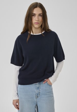 Marineblauer Strickpullover mit kurzen Ärmeln und rundem Ausschnitt, darüber getragen ein weißes Langarmshirt, kombiniert mit hellblauen Jeans.