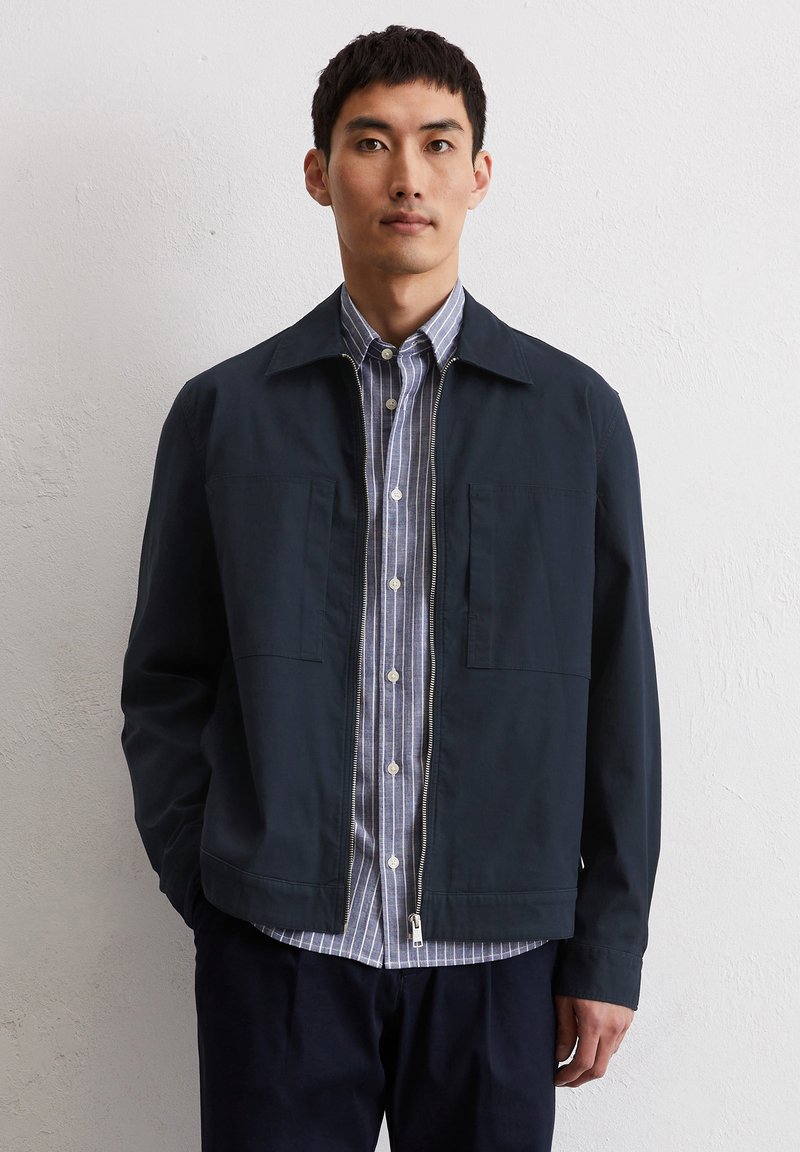 Navy jacket with a flat collar and two front pockets, worn over a light blue and white striped button-up shirt, displayed against a plain background.
