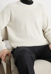 Krémový žebrovaný svetr s kulatým výstřihem, nošený přes černý rolák, spárovaný s černými kalhotami, sedící na světlé židli.