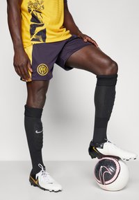 Nike voetbaloutfit met een gele top met een grafisch patroon, marineblauwe shorts, zwarte sokken en wit/rode voetbalschoenen op een voetbal.