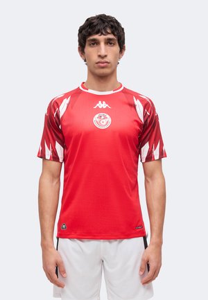 Man met een rood sportshirt met wit en donkerrood patroon op de mouwen en witte korte broek, staand tegen een effen achtergrond.