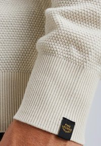 Gebreide trui detail in off-white met een gestructureerde oppervlakte. Ribbelmanchetten en een zwart label met "PME LEGEND" in gouden accenten.