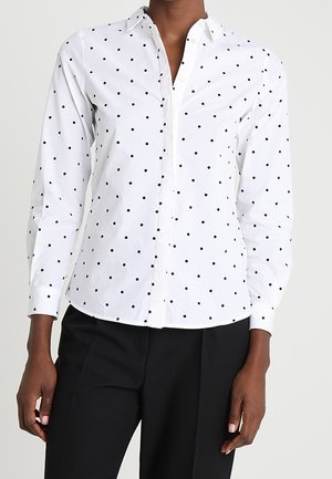 Chemise blanche à manches longues avec des pois noirs, portée avec un pantalon noir, montrée du cou aux hanches.