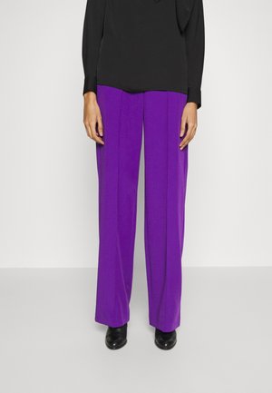 Person wearing purple wide-leg trousers, black long-sleeve top, and black shoes standing against a plain background.