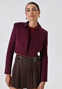 Giacca cropped bordeaux con colletto alto e chiusura a zip, abbinata a una gonna plissettata marrone e una cintura a due fibbie. Tessuto liscio e strutturato.