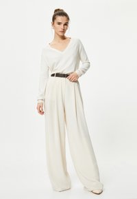 Biały sweter w serek, zestawiony z kremowymi spodniami o szerokich nogawkach, związany w talii czarnym paskiem, gładka faktura, minimalistyczny design.