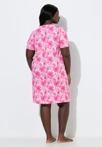 Mujer de pie descalza, vistiendo un vestido rosa con flores de manga corta hasta la rodilla, de espaldas sobre un fondo blanco liso.