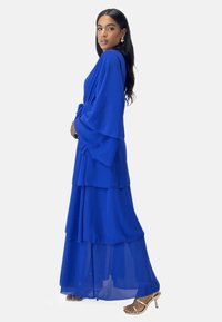 Blå, lagdelt chiffon maxi-kjole med brede ærmer, indsnøret talje og gennemsigtigt lag. Kombineret med gyldne hæle sandaler og hængende øreringe.