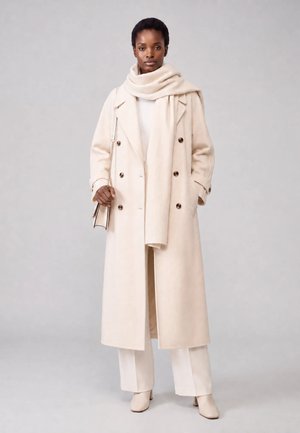 Frau steht, trägt einen langen cremefarbenen Mantel, passenden breiten Schal, weiße Hose und cremefarbene Stiefel, hält eine kleine beige Handtasche mit Kettenriemen.
