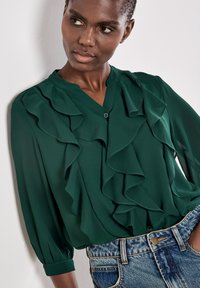 Blusa verde leggera con scollo a V, decorata con rouches a cascata e maniche a tre quarti, abbinata a pantaloni shorts in denim.