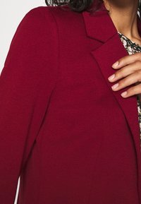 Femme portant un blazer rouge profond sur un haut à motifs floraux, tenant le revers du blazer de la main droite.