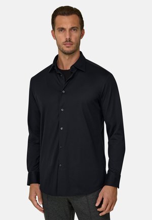 Homme portant une chemise noire cintrée à manches longues boutonnée et un pantalon gris, debout devant un fond clair uni.