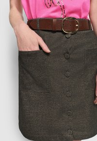 Camisa rosa metida dentro de una falda gris oscuro abotonada con grandes bolsillos y un cinturón de textura marrón, con una mano descansando en el bolsillo.