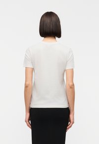 Γυναίκα με ίσιο σκούρο καρέ μαλλί, ντυμένη με απλό λευκό κοντομάνικο μπλουζάκι και μαύρη φούστα, στέκεται με την πλάτη.