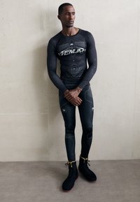 Sort atletisk langærmet skjorte og leggings med geometriske mønstre og hvide accenter, parret med sorte højhælede sko med gule detaljer.
