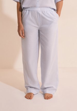 Persoon draagt lichtblauwe gestructureerde losse broek met elastische tailleband en is blootsvoets, staand tegen een beige achtergrond.