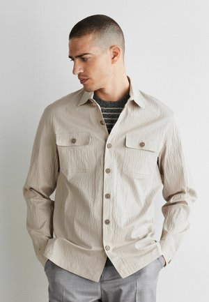 Mand med kort hår iført en beige tekstureret skjorte med knapper over en stribet mørkegrå sweater, står med hænderne i lommerne.