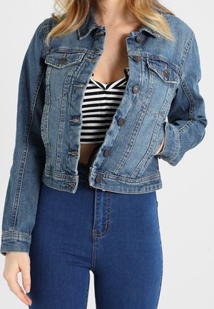 Femme portant une veste en jean bleue par-dessus un crop top rayé noir et blanc, associée à un jean bleu taille haute, une main dans la poche de la veste.