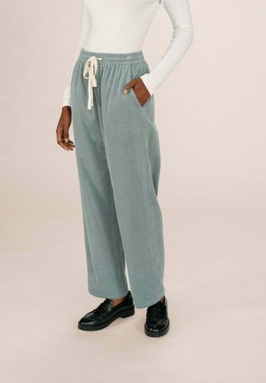 Personne portant un pantalon en velours côtelé large bleu clair avec un cordon blanc, un haut à manches longues blanc et des mocassins en cuir noirs, debout.