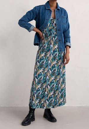 Person, die ein langes florales Kleid mit türkis-, beige- und weißfarbenen Mustern, eine blaue Jeansjacke und schwarze Schnürstiefel trägt, steht drinnen.