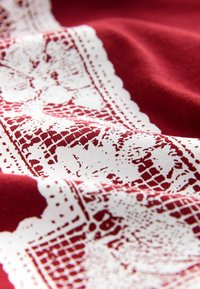 Raudona medžiaga su balto nėrinių raštu ir sudėtingomis detalėmis. Tekstūra atrodo lygi su švelniu blizgesiu, o kraštas yra banguotas.