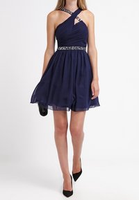 Marinblå chiffongklänning med figurnära bodice, rynkade detaljer och ett juvelbesatt midjeband. Har korsade axelremmar och en utsvängd kjol.