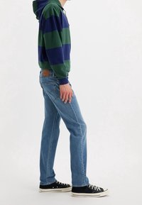 Sudadera con capucha de rayas verdes y marinas, combinada con jeans de mezclilla azul claro y zapatillas negras con suelas blancas.
