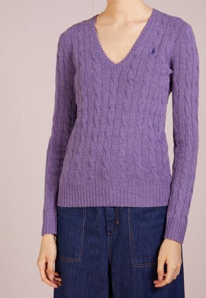 Femme portant un pull violet en maille torsadée à col en V et un pantalon en jean bleu foncé, debout devant un fond clair et neutre.