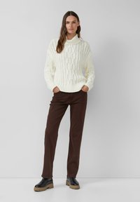 Pull col roulé en tricot épais crème, associé à un pantalon droit marron et des bottes à semelle épaisse noires. Détails de tricot texturé visibles.