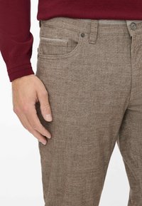 Braune, strukturierte Hose mit subtilen Mustern, ausgestattet mit fünf Taschen und im Slim-Fit-Schnitt, präsentiert neben einer Hand, die auf dem Stoff ruht.