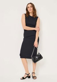 Vestido midi negro sin mangas con un diseño ajustado, textura suave, combinado con sandalias de tacón negras y un pequeño bolso negro.