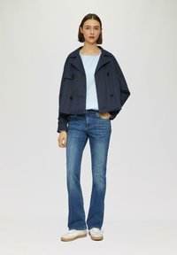 Giacca corta blu navy con bottoni doppi, top a righe blu chiaro, jeans aderenti e sneakers bianche con suole beige.
