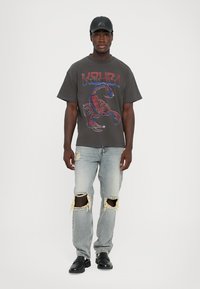 Tricou grafic închis, cu un design de scorpion roșu, asociat cu blugi deschiși la culoare, deteriorați, având rupturi la genunchi, și încălțăminte neagră. Accesorii: șapcă neagră.