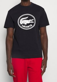 Czarny bawełniany t-shirt z białym, okrągłym logo Lacoste z wizerunkiem aligatora. Krótkie rękawy, okrągły dekolt, luźny krój.