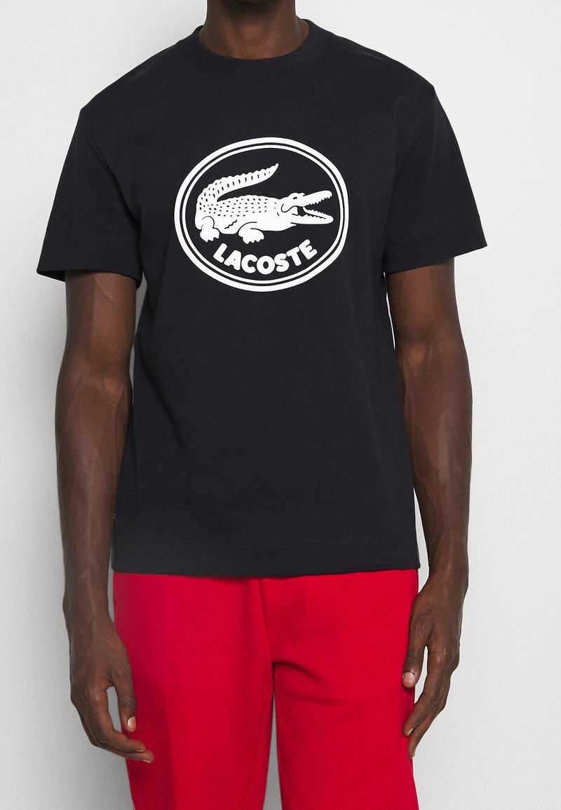Czarny bawełniany t-shirt z białym, okrągłym logo Lacoste z wizerunkiem aligatora. Krótkie rękawy, okrągły dekolt, luźny krój.