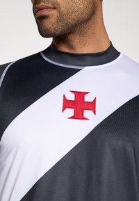 Atletska majica z zasnovo sivih in belih barvnih blokov, z rdečim vezenim križem na levem prsnem delu in visokim izrezom na vratu.