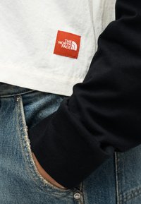 Λευκό πουκάμισο με κόκκινη ετικέτα λογότυπου The North Face, μαύρο μανίκι και μπλε τζιν από denim, με το χέρι στην τσέπη ορατό.