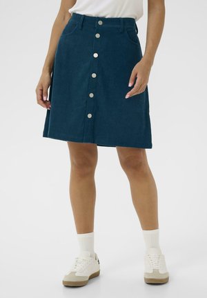 Jupe en velours côtelé de couleur bleu canard, de forme évasée, avec une fermeture à boutons sur le devant, deux poches latérales et une finition texturée. Associée à des baskets blanches.