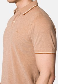 Jasnobrązowa koszulka polo z teksturowanej tkaniny, z kontrastowym kołnierzykiem w cienkie paski i małym logo na klatce piersiowej.