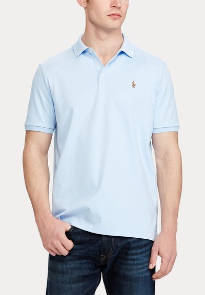 Mies, joka pukeutunut vaaleansiniseen lyhythihaiseen poolopaitaan, jossa on pieni kirjailtu logo rinnassa, ja tummansinisiin farkkuihin, käsi taskussa.