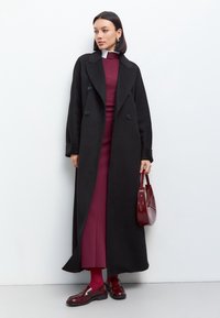 Palton lung negru cu nasturi mari, purtat peste o rochie burgundy și colanți asortați. Accesoriile includ o geantă burgundy și pantofi luciosi.