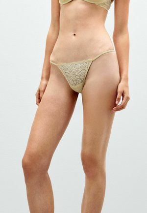 Kvinde iført beige blondeundertøj, der viser overkrop og øvre ben mod en ensartet lys baggrund.