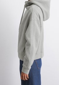 Szara bluza z kapturem, wykonana z miękkiego materiału, z regulowanym kapturem, kieszenią kangurką i ściągaczami na rękawach. Połączona z niebieskimi dżinsami.