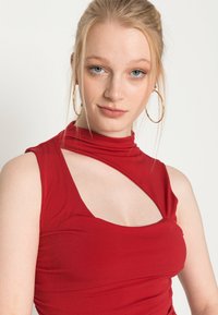 Top sem mangas vermelho em tecido suave com um decote alto e detalhe em recorte lateral, apresentando um design ajustado e uma textura lisa.