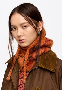 Chaqueta de pana marrón con un cuello ancho, superpuesta a una blusa naranja con un estampado. La modelo lleva una bufanda de felpa naranja con detalles en negro.