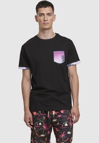 Juoda medvilninė trumpomis rankovėmis marškinėliai su purpuriniu kišenės atspaudu ir kontrastingomis raštuotomis rankogaliais. Modelis dėvi spalvotus raštuotus šortus.