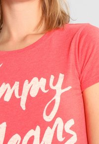 Korallinen puuvillapaita, jossa on lyhyet hihat, avara kaula-aukko ja valkoinen teksti, joka lukee "Iloisia Unelmia" leikkisällä fontilla.